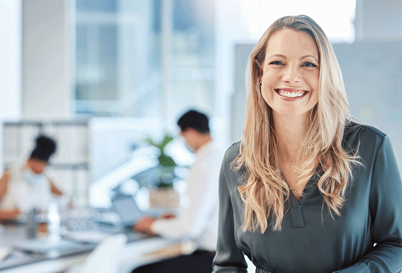 Qualität bei 3PAGEN Qualität bei 3PAGEN - eine Frau lächelt in die Kamera, im Hintergrund ist ein Büro zu sehen, wo verschwommen Kollegen Produkte begutachten